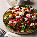 A large plate of salad with roasted sweet potatoes, broccoli, arugula, pomegranate, and goat cheese.