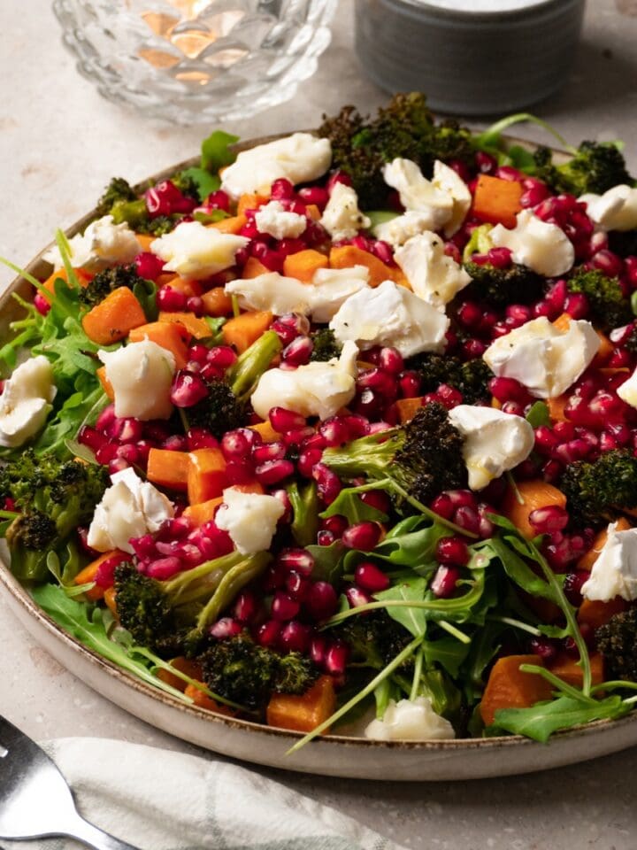A large plate of salad with roasted sweet potatoes, broccoli, arugula, pomegranate, and goat cheese.
