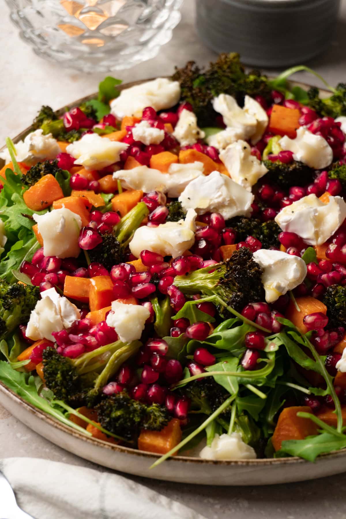A large plate of salad with roasted sweet potatoes, broccoli, arugula, pomegranate, and goat cheese.