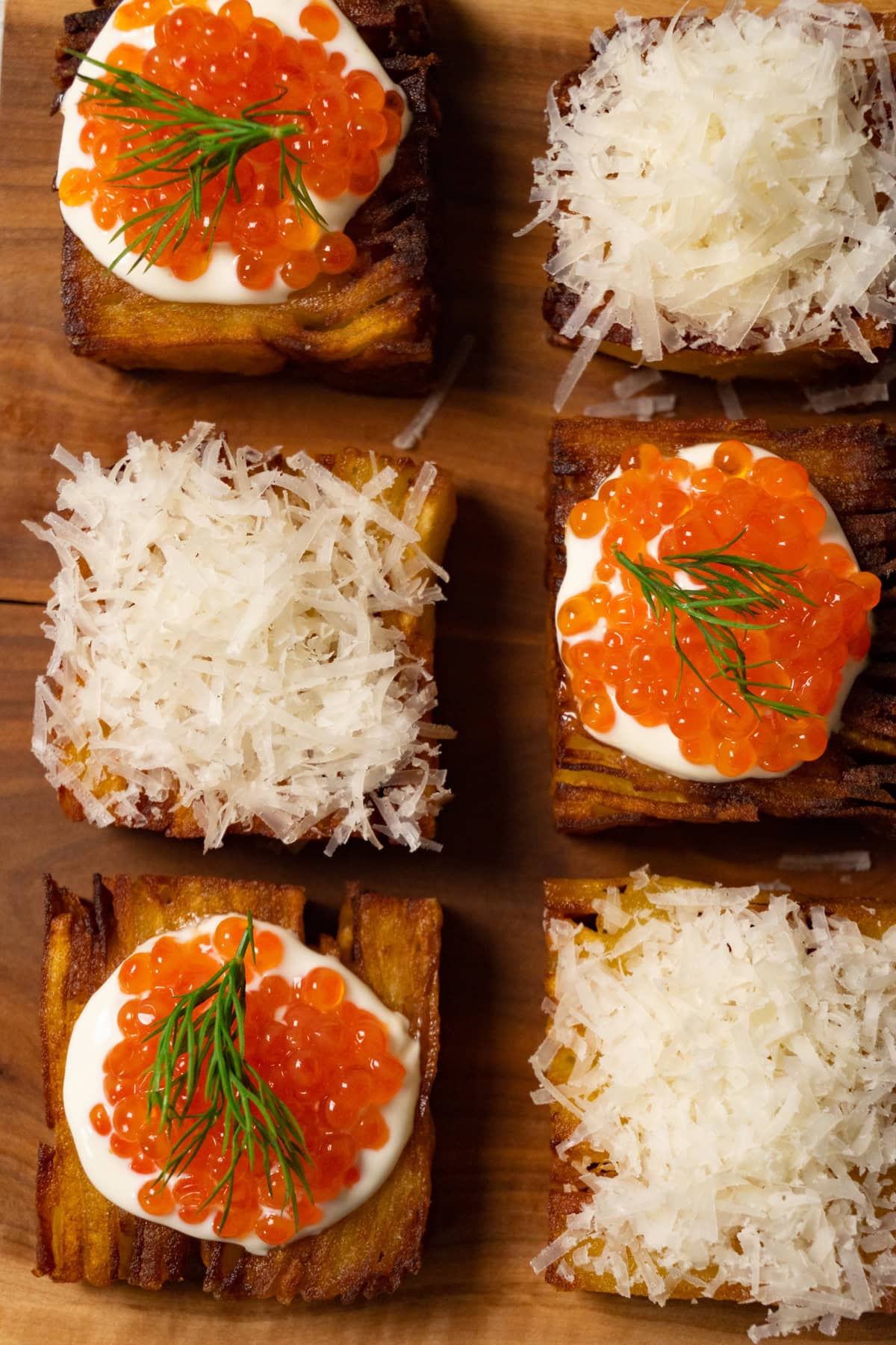 Six pieces of potato pavé are arranged on a wooden board, some topped with shredded Parmesan and others with sour cream and red caviar.