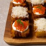 Six pieces of potato pavé are arranged on a wooden board, some topped with shredded Parmesan and others with sour cream and red caviar.