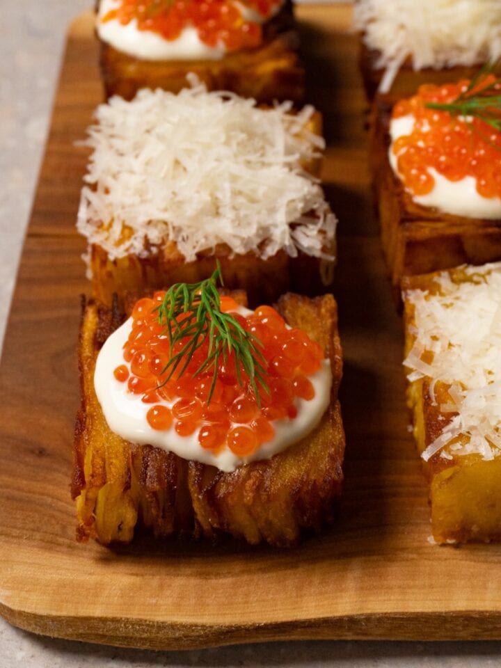 Six pieces of potato pavé are arranged on a wooden board, some topped with shredded Parmesan and others with sour cream and red caviar.