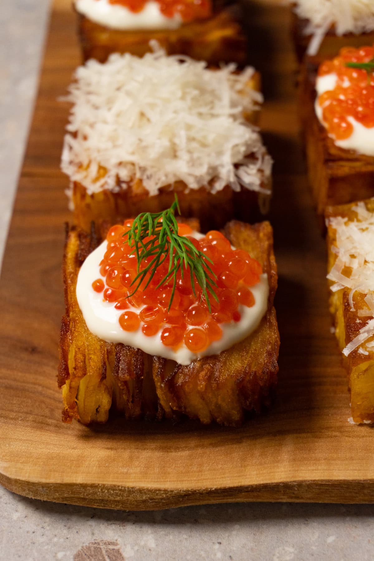 Six pieces of potato pavé are arranged on a wooden board, some topped with shredded Parmesan and others with sour cream and red caviar.
