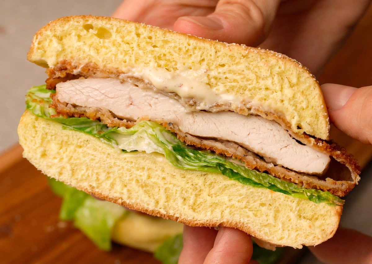 Hands are holding a sandwich cut in half with crispy chicken, salad, and white sauce.