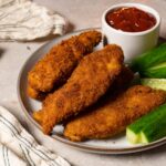 Chicken tenders coated in a crispy crust are served on a plate with fresh cucumbers and ketchup.