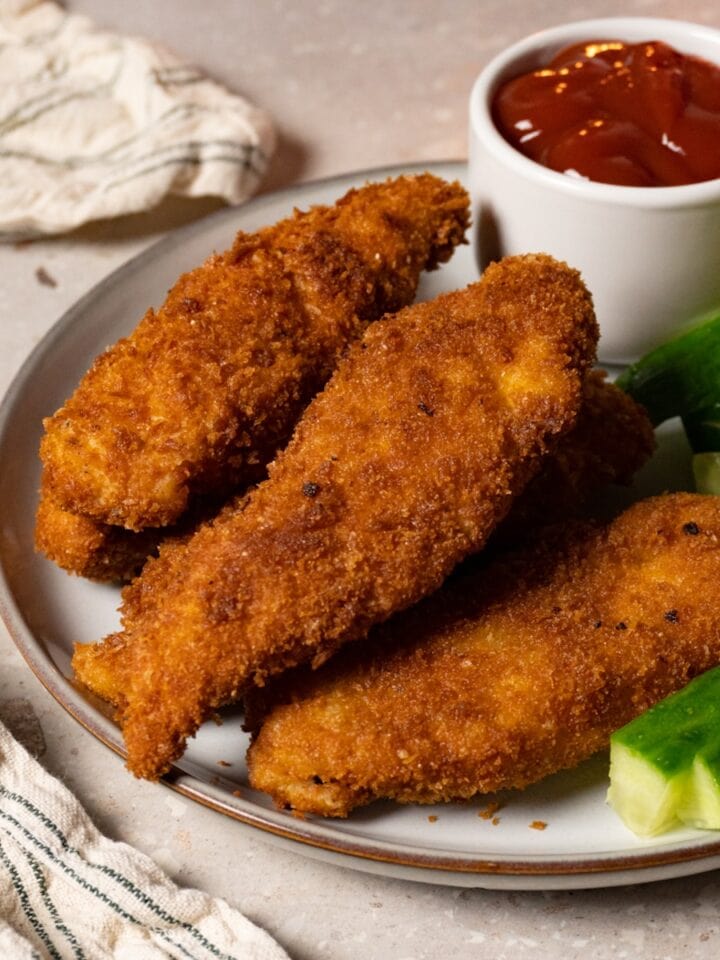 Chicken tenders coated in a crispy crust are served on a plate with fresh cucumbers and ketchup.