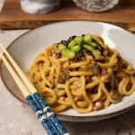 A bowl of udon noodles in a creamy sauce with mushrooms and pancetta, topped with nori and green onions.