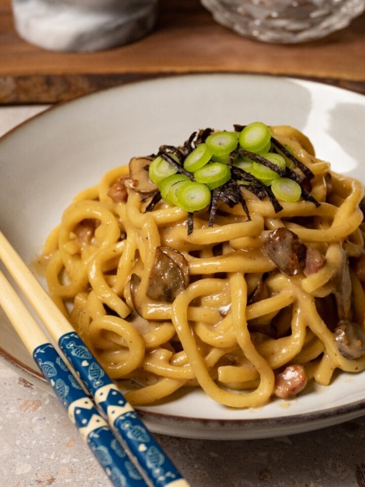 A bowl of udon noodles in a creamy sauce with mushrooms and pancetta, topped with nori and green onions.