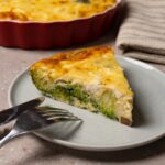 A slice of crustless quiche with broccoli, mushrooms and cheese on a plate with a fork and knife.