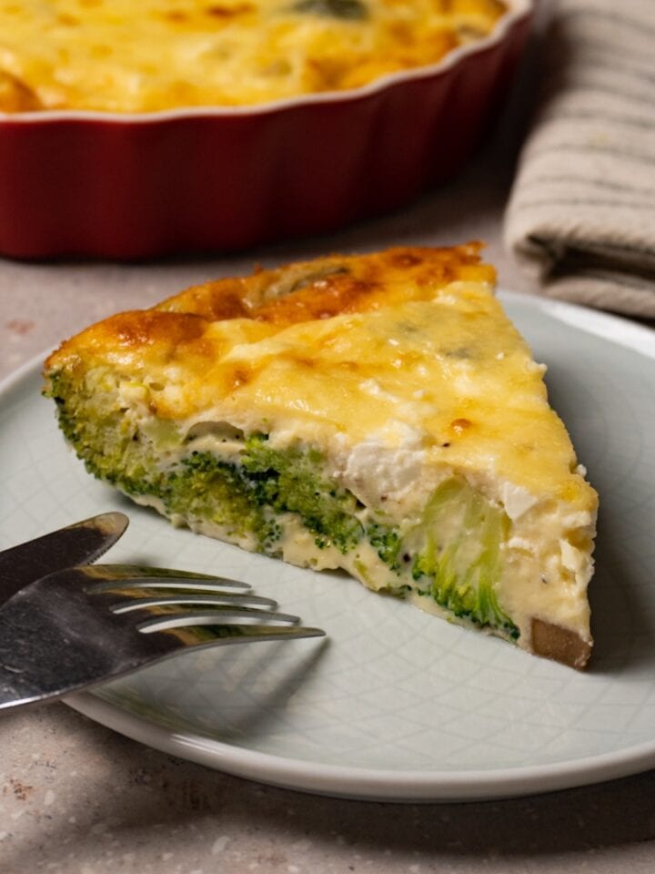 A slice of crustless quiche with broccoli, mushrooms and cheese on a plate with a fork and knife.