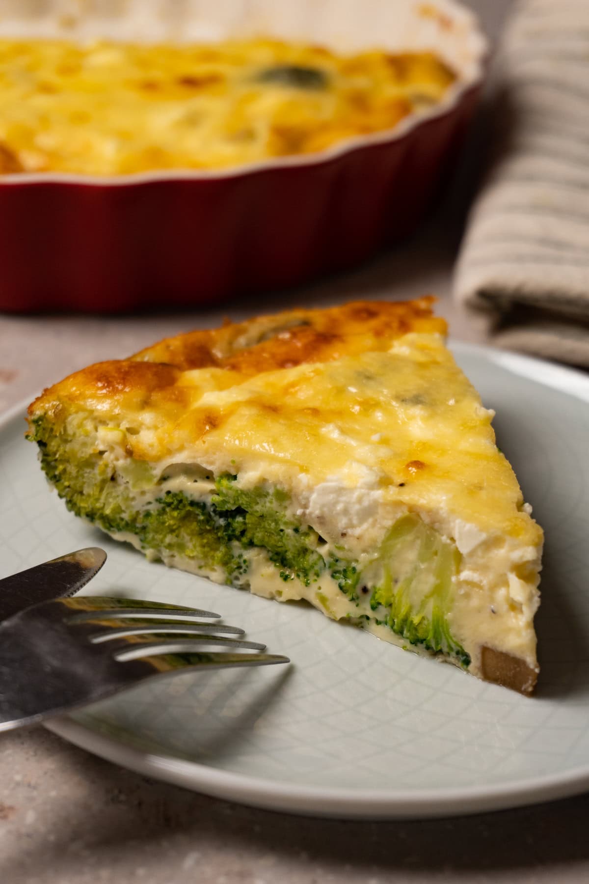 A slice of crustless quiche with broccoli, mushrooms and cheese on a plate with a fork and knife.