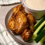 Chicken wings covered in a shiny sauce are served on a plate with fresh cucumbers and a bowl of white sauce.