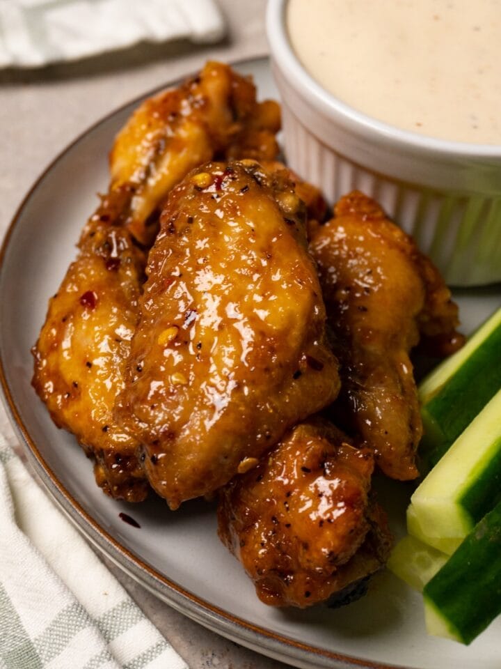 Chicken wings covered in a shiny sauce are served on a plate with fresh cucumbers and a bowl of white sauce.