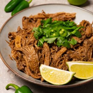 A bowl of pulled pork garnished with fresh cilantro, jalapeños, and lime wedges.