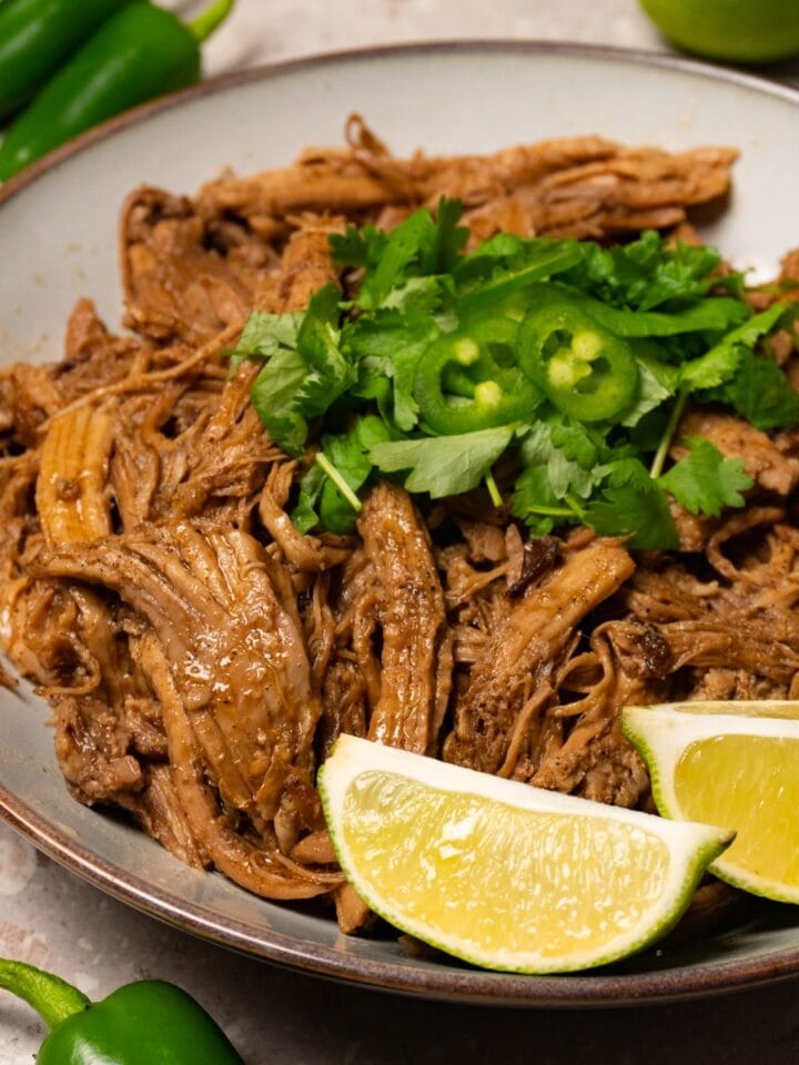A bowl of pulled pork garnished with fresh cilantro, jalapeños, and lime wedges.