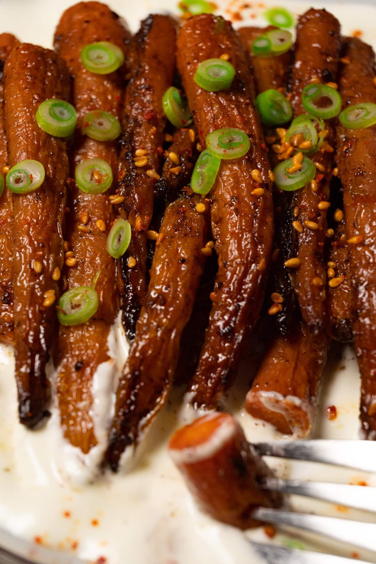 Roasted carrots topped with toasted sesame seeds and green onions, served with white sauce on a plate. One piece has been taken from a carrot.