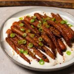 Roasted carrots topped with toasted sesame seeds and green onions, served with white sauce on a plate.