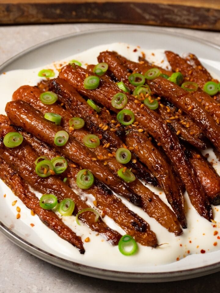 Roasted carrots topped with toasted sesame seeds and green onions, served with white sauce on a plate.