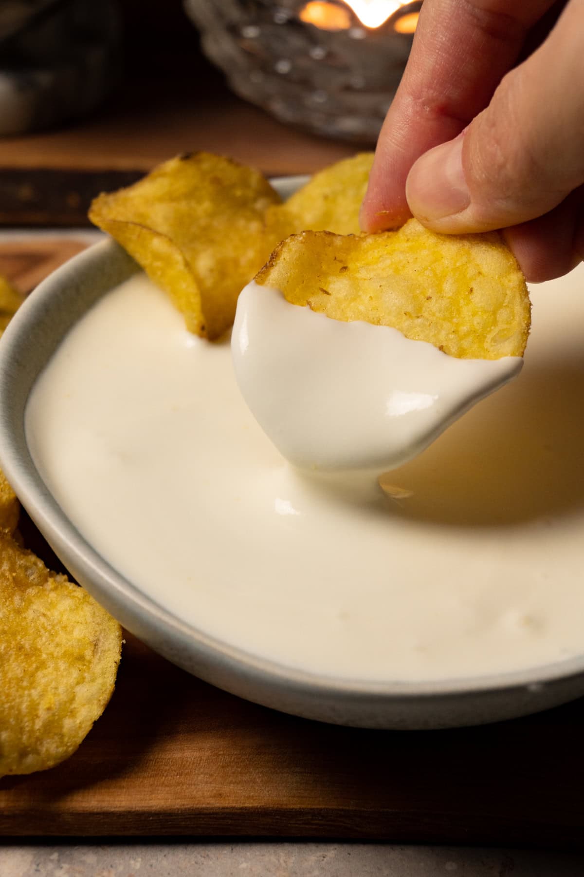 A hand is dipping a potato chip into a bowl of white dipping sauce.