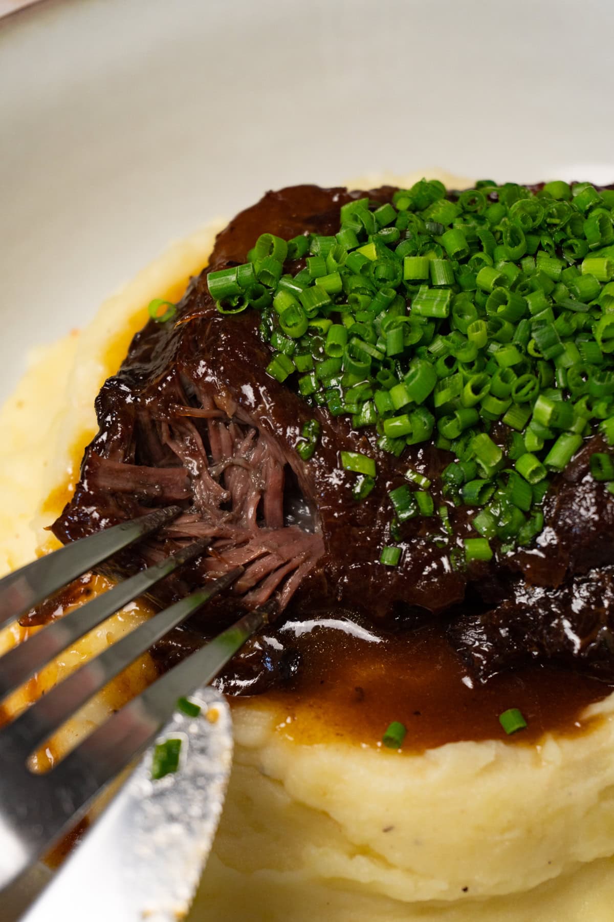 A fork is digging into braised beef cheek served on top of mashed potatoes and topped with finely chopped chives.