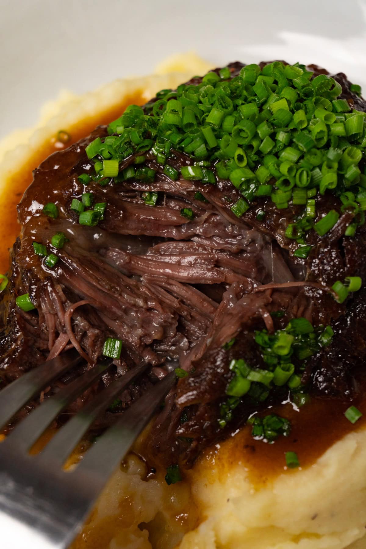 A fork is digging into braised beef cheek served on top of mashed potatoes and topped with finely chopped chives.