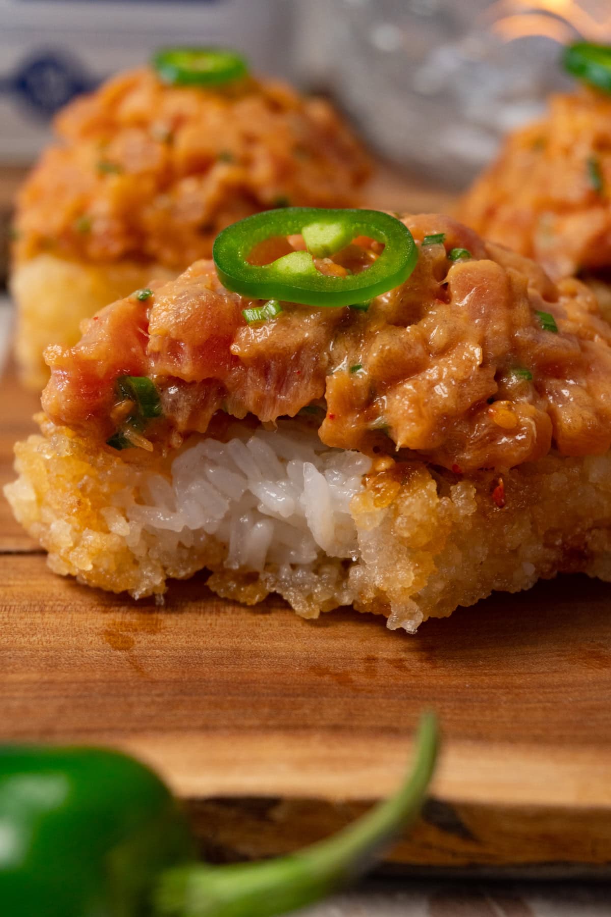 Crispy rice cakes topped with spicy tuna and sliced fresh jalapeños are served on a wooden board. One of the cakes has a bite taken out of it.