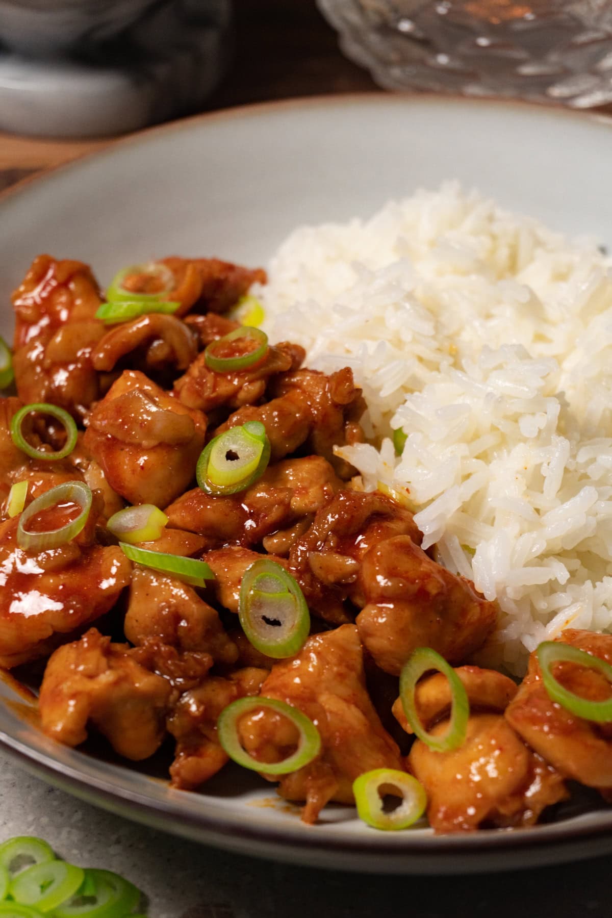 A bowl of bite-sized chicken covered in red sauce, topped with sliced green onions, with white rice on the side.