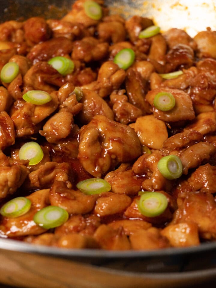 A skillet with bite-sized chicken covered in red sauce and topped with sliced green onions.