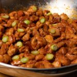 A skillet with bite-sized chicken covered in red sauce and topped with sliced green onions.