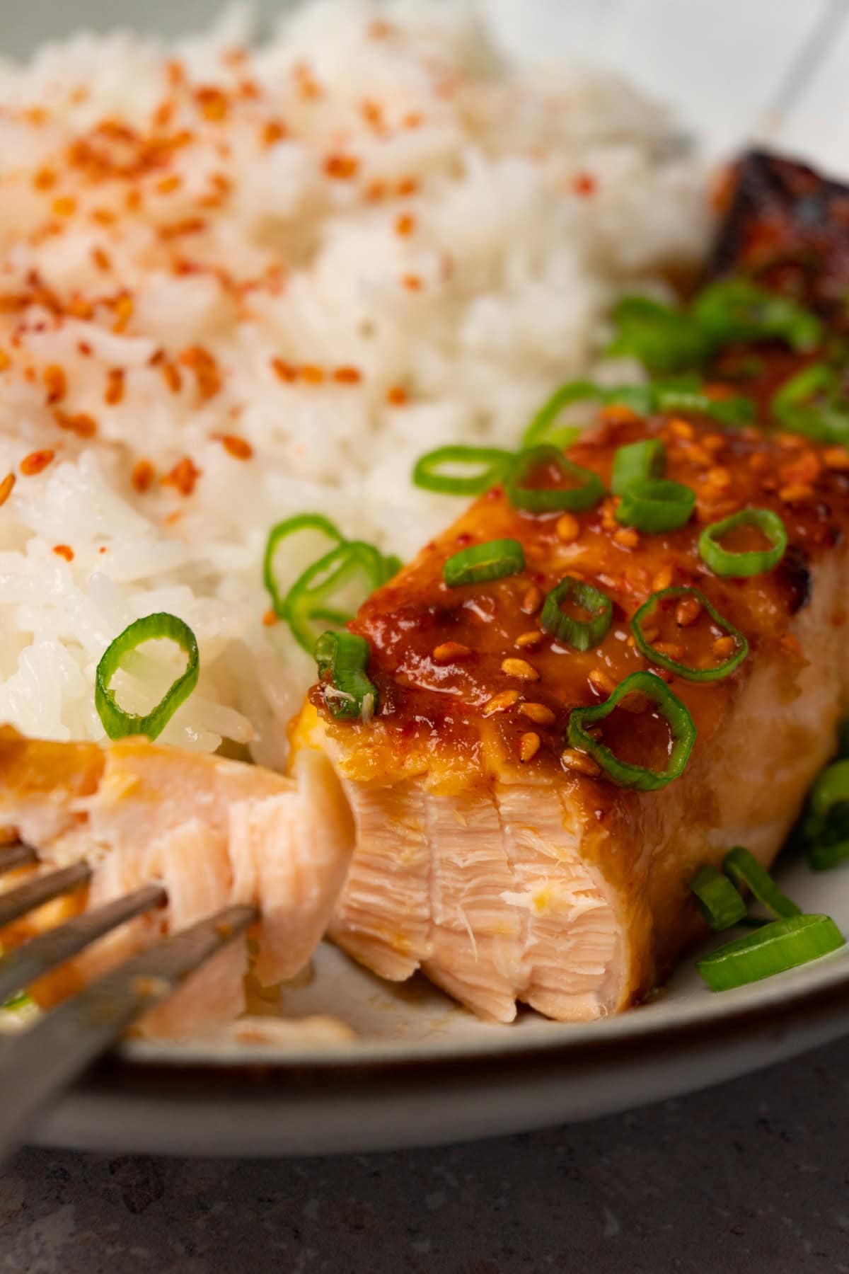 Glazed salmon fillets topped with sesame seeds and green onions in a bowl with jasmine rice on the side, with one bite taken from the fillet.