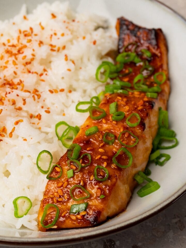 Glazed salmon fillets topped with sesame seeds and green onions in a bowl with jasmine rice on the side.