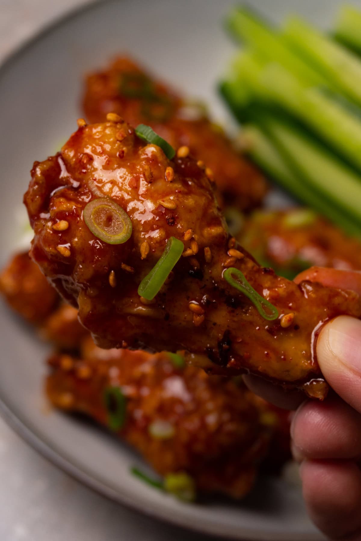 A hand is holding chicken wings covered in red sauce and topped with toasted sesame seeds and freshly chopped onion.