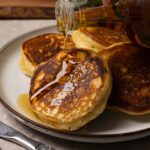 Pancakes served on a plate are being topped with maple syrup.