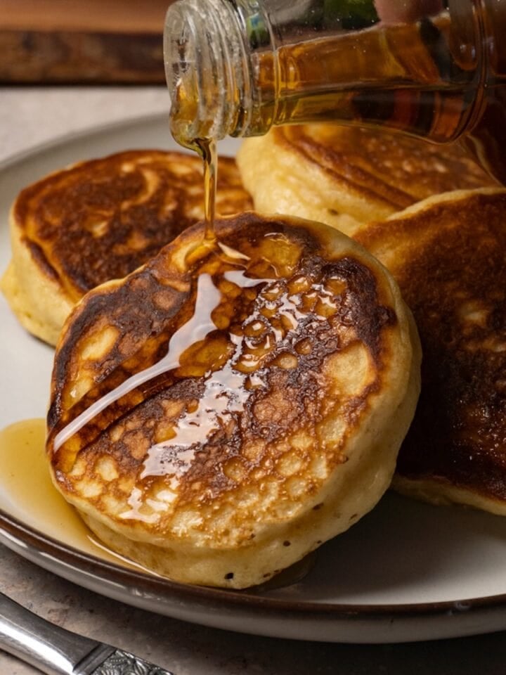 Pancakes served on a plate are being topped with maple syrup.